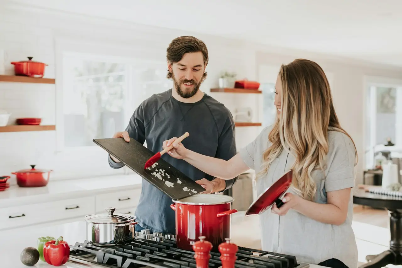 Two people cooking together at a home stove, lifting the lid on a large red pot.